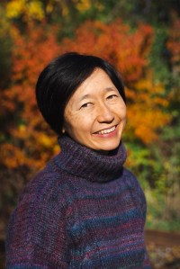 Smiling middle-aged Asian cisgender woman with short hair wearing a purple wool sweater against orange and green foliage.