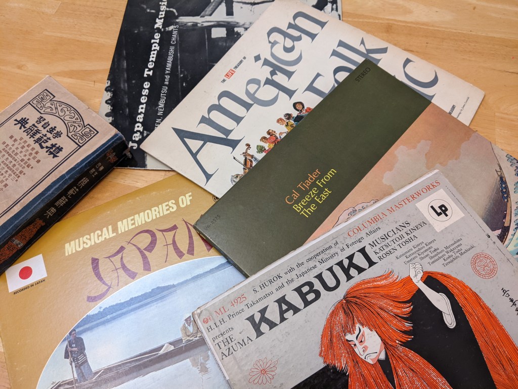 Blonde wood table top with phonograph records of Japanese music marketed for an English-speaking audience, phonograph record of American folk music, and an old Japanese character dictionary.