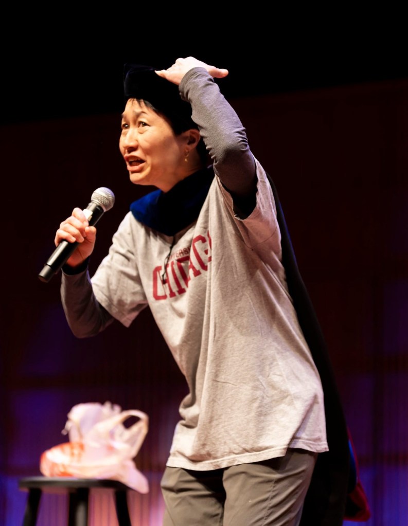 Asian woman with short hair in black PhD tam and dark blue hood wearing gray University of Chicago T-shirt, black long sleeved shirt, and olive green pants holding a microphone in her right hand and motioning toward her tam with the left, with a humorously anguished expression. In the back is a stool with a white and orange Home Depot plastic bag.