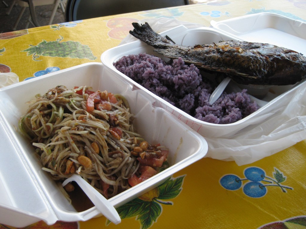 Papaya salad, fried tilapia, and purple rice with plastic utensils in styrofoam containers.